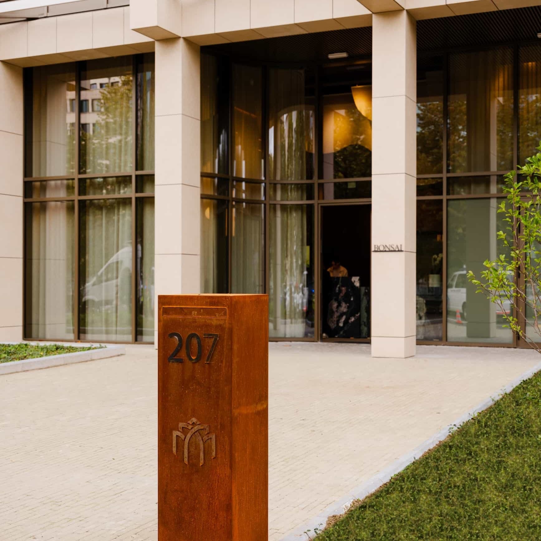 The front entrance of the Bonsai club in Brussels, with floor to ceiling glass, cream stone pillars and the number 207 on a corten steel bollard