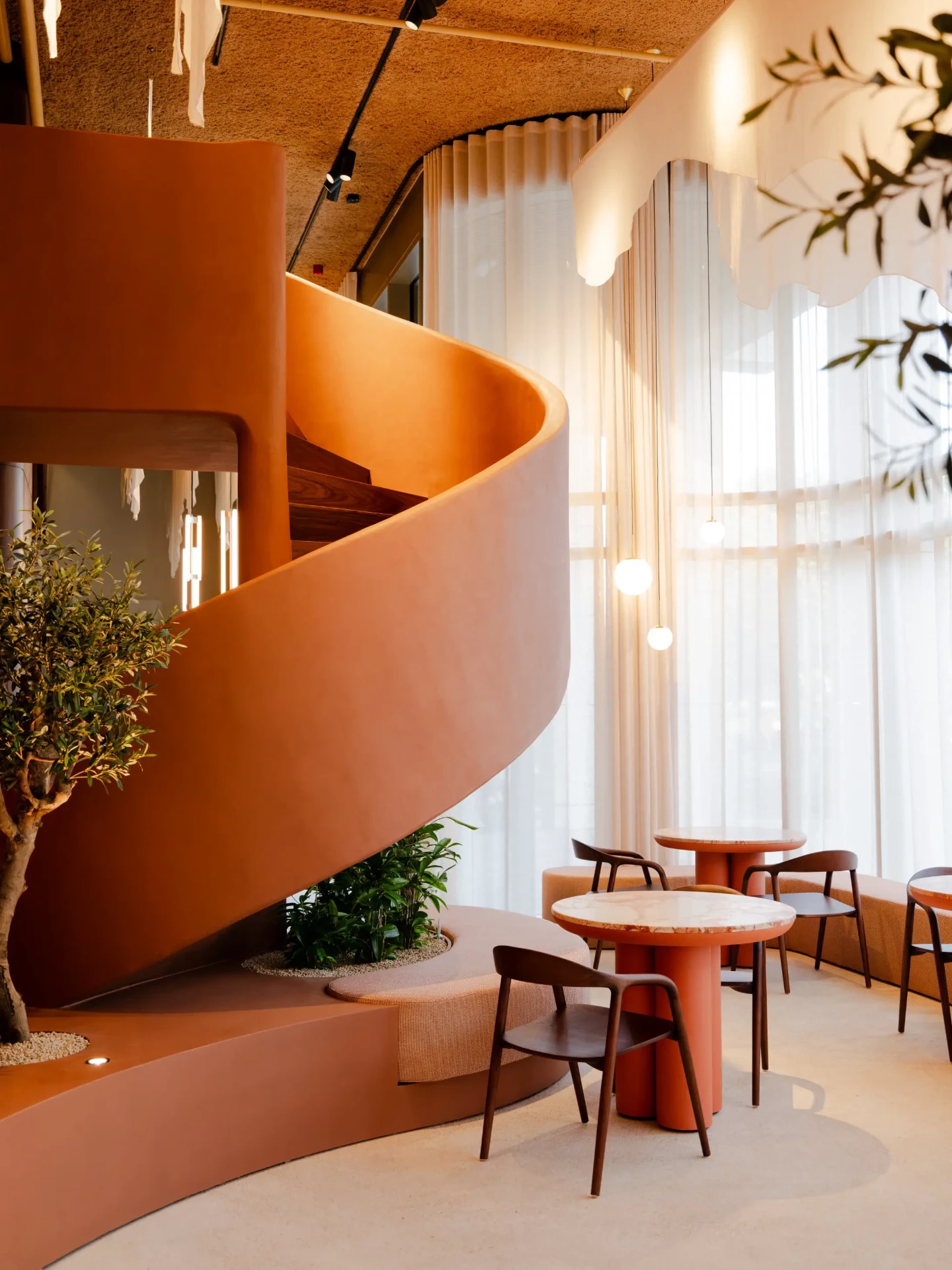 Bonsai lounge with a terracotta spiral staircase, an olive tree and marble tables by sheer curtains in soft daylight